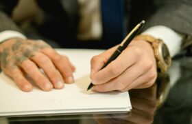 A close-up view of a businessman with tattoos signing a document at a desk.