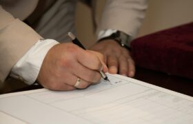 A close-up of a man signing a document, showcasing a wedding ring and pen.