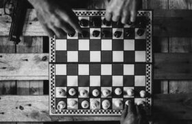 Black and white chessboard setup with hands moving pieces on a wooden table alongside a gun.