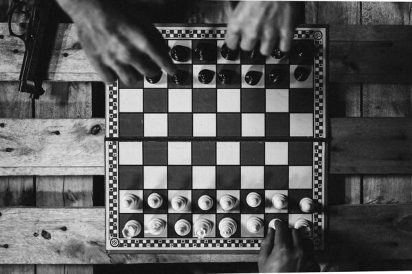 Black and white chessboard setup with hands moving pieces on a wooden table alongside a gun.